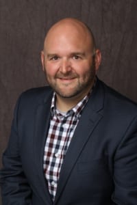 James Oltman smiles while wearing a checkered shirt under a dark jacket