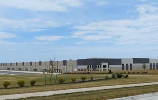 Clouds dot a blue sky over a newly built warehouse at Logistics Park Kansas City. The warehouse is set back from the road behind newly planted trees and shrubs. The building itself is primarily a creamy beige with articulations of slate gray and white trim.